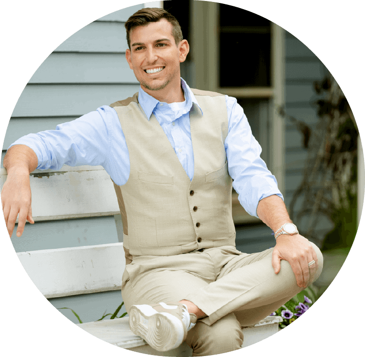 Matt Fraser, dressed in a beige vest, light blue shirt, and beige pants, sits smiling on a white bench in front of a house.